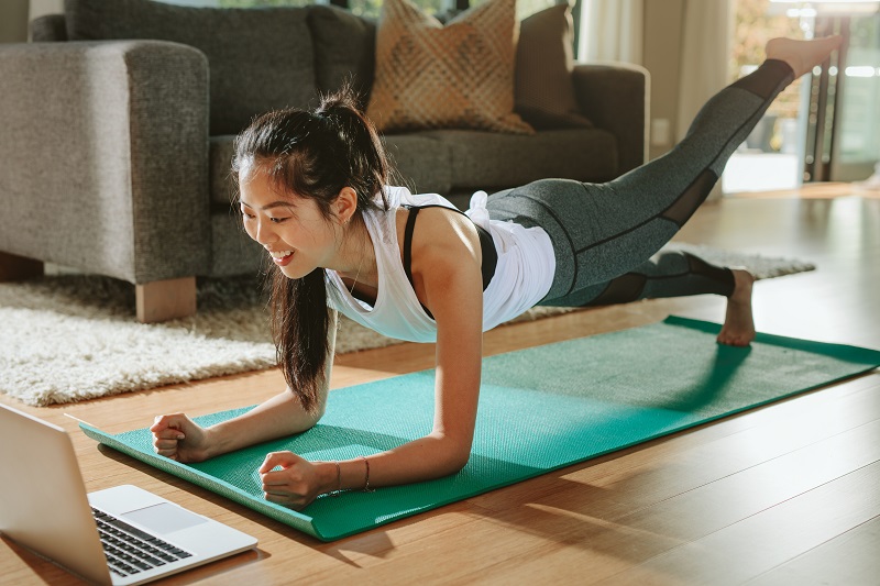 Smiling woman exercising at home and watching training videos on laptop. Chinese female doing planks with a leg outstretched and looking at laptop.