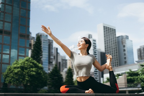 Athletic young Asian woman practicing yoga outdoors in city park against urban cityscape in the morning
