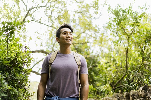 A photo of confident hiker standing against trees. Smiling young men is wearing casuals. He is in forest.